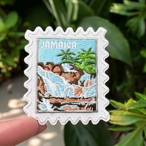 May include: White embroidered patch with a blue and green image of a waterfall and palm trees. The patch has a white border with a scalloped edge and the word "Jamaica" printed in black.
