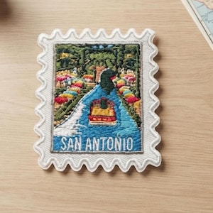 May include: A rectangular embroidered patch shaped like a postage stamp with scalloped edges. The design features a colorful river scene with a boat, lush greenery, and flowers. The text SAN ANTONIO is stitched in white at the bottom of the patch.
