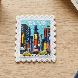 May include: A rectangular embroidered patch shaped like a postage stamp with a scalloped white border. The design features a colourful New York City skyline with tall buildings, yellow taxis, and the text NEW YORK in white letters at the top.