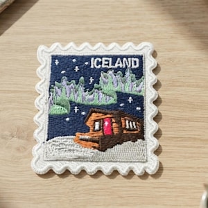 May include: A rectangular embroidered patch shaped like a postage stamp with a white scalloped border. The design features a brown cabin with a bright red door, a snowy foreground, and green aurora borealis lights against a dark blue sky with the text ICELAND.