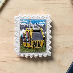 May include: A rectangular embroidered patch shaped like a postage stamp with a scalloped white border. The design features a yellow gondola car with the word VAIL in black letters, set against a backdrop of blue mountains and green trees.