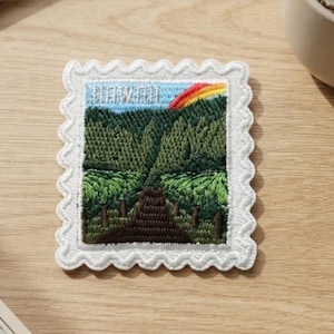 May include: A rectangular embroidered patch with a scalloped white border, featuring a landscape scene of a dark brown path leading through dense green forests toward a blue sky with a colorful rainbow and the word HAWAII stitched in white.