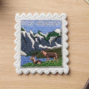 May include: A rectangular embroidered patch shaped like a postage stamp with a white scalloped border. The design features the text ROCKY MOUNTAINS above a landscape of blue mountains with white snow, green hills, and two brown deer in the foreground.