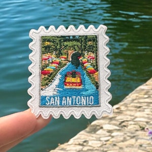 May include: White embroidered patch featuring a colourful image of a boat on a river with trees and flowers in the background. The text "SAN ANTONIO" is printed in black at the bottom of the patch.