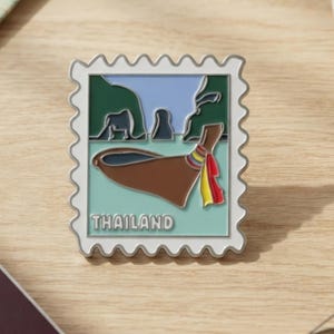 May include: A rectangular enamel pin shaped like a postage stamp featuring a brown long-tail boat with a colorful fabric wrap on the prow, set against a light blue water and dark green cliff background with the word THAILAND printed in white.