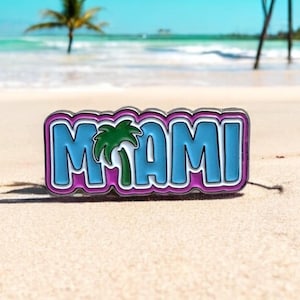 Peut inclure: Une épingle en émail bleu et rose avec le mot "MIAMI" en lettres blanches et une icône de palmier vert. L'épingle est sur une plage de sable blanc avec un arrière-plan flou d'eau bleue et de palmiers.