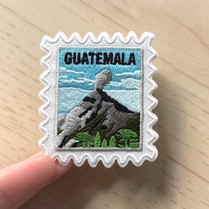 May include: A rectangular embroidered patch shaped like a postage stamp with a white scalloped border. The design features a mountain landscape with a smoking volcano, green trees, and a blue sky. The word GUATEMALA is stitched in black letters.
