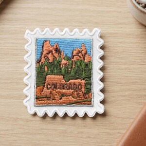 May include: A rectangular embroidered patch shaped like a postage stamp with a white scalloped border. The design features a landscape of orange rock formations, green pine trees, and a blue sky with the word COLORADO stitched in brown thread.