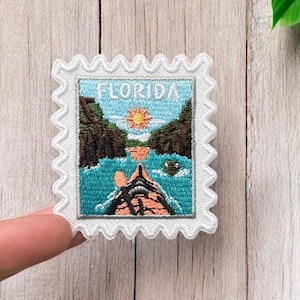 Könnte beinhalten: Weißer gestickter Aufnäher mit einem blauen und grünen Bild eines Kajaks in einem Fluss mit dem Text "FLORIDA" in Weiß.