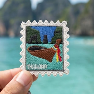 Puede incluir: Un parche bordado cuadrado con forma de sello postal y borde blanco festoneado. El diseño muestra una barca tradicional marrón con cintas rojas y amarillas sobre agua turquesa, acantilados verde oscuro de fondo y la palabra THAILAND.