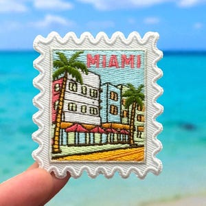 May include: Embroidered patch in the shape of a postage stamp featuring a Miami scene with palm trees and buildings. The word "MIAMI" is displayed in pink. The patch has a white border with a scalloped edge.