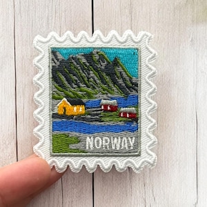 Könnte beinhalten: Gestickter Aufnäher mit einer norwegischen Landschaft, gestaltet wie eine Briefmarke. Er zeigt ein gelbes Haus, rote Häuser, Berge und das Wort "NORWAY". Der Aufnäher hat einen weißen, gezackten Rand.