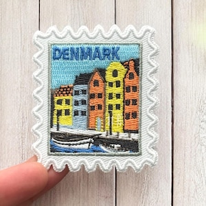 May include: Embroidered patch in the shape of a postage stamp featuring the word "DENMARK" in blue above a colorful illustration of buildings and a boat. The patch has a white, scalloped border.