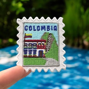Può includere: Una toppa ricamata rettangolare a forma di francobollo con un bordo bianco smerlato. Il disegno raffigura un edificio con una bandiera colombiana, colline verdi e un sentiero, con la parola COLOMBIA ricamata in blu nella parte superiore.