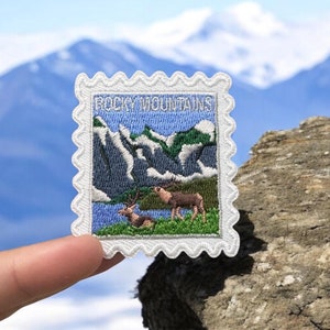 May include: White embroidered patch with a blue and green mountain scene and two deer. The patch has a scalloped edge and says "Rocky Mountains" in white text.