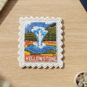 May include: A rectangular embroidered patch shaped like a postage stamp with a scalloped white border. The design features a blue and white geyser erupting against a green forest and blue sky, with the word YELLOWSTONE printed in white letters.
