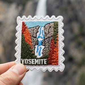 May include: A square embroidered patch with a scalloped white border featuring a landscape of a waterfall cascading down rocky cliffs, surrounded by green and orange trees, with the word YOSEMITE stitched in white at the bottom of the design.