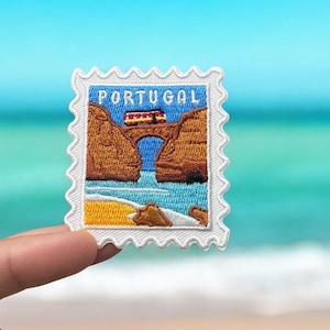 Op de afbeelding: Witte geborduurde patch met een stempelontwerp met een brug over een waterpartij met een geel strand en een blauwe lucht. De tekst "PORTUGAL" is in wit op de patch gedrukt.