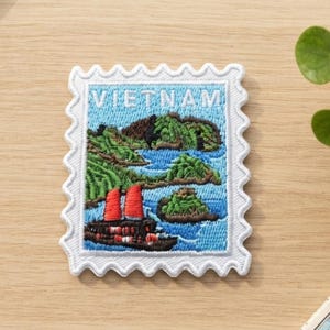 Könnte beinhalten: Ein rechteckiger Stickaufnäher in Form einer Briefmarke mit weißem, gewelltem Rand. Das Motiv zeigt ein blaues Meer, grüne felsige Inseln und ein dunkles Boot mit zwei roten Segeln. Das Wort VIETNAM ist oben in weißer Schrift eingestickt.