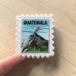 May include: Embroidered patch in the shape of a postage stamp featuring a mountain landscape. The patch has a white border with a scalloped edge. The word "GUATEMALA" is embroidered in blue letters across the top of the patch.
