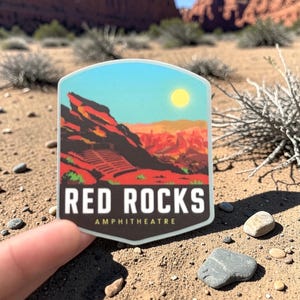 May include: A die-cut sticker featuring a stylized illustration of the Red Rocks Amphitheatre with red rock formations, a blue sky, and a yellow sun. The text RED ROCKS AMPHITHEATRE is printed in white and yellow on a dark brown background at the base.