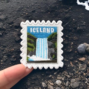 May include: A rectangular embroidered patch with a scalloped white border, featuring a landscape illustration of a waterfall between green hills and rocky terrain, with the word ICELAND in white capital letters against a blue sky background.