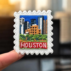 May include: Embroidered patch in the shape of a postage stamp featuring a cityscape with the word "HOUSTON" in red letters. The patch has a white border with a scalloped edge, and the cityscape includes buildings and green foliage.