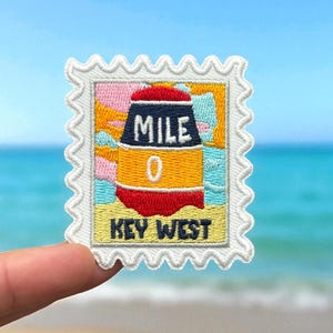 May include: Embroidered patch featuring the iconic Mile 0 buoy of Key West, Florida, in a postage stamp design. The buoy is depicted with a red top, black and yellow body, and the text "MILE 0 KEY WEST". The background has colorful abstract shapes.