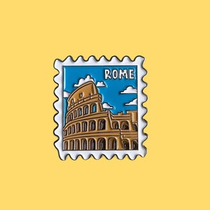 May include: A white enamel pin with a blue and brown image of the Colosseum in Rome. The pin has a white border and the word "ROME" in white letters.