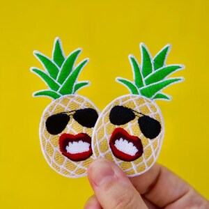 May include: Two yellow pineapple embroidered patches with smiling faces wearing sunglasses. The pineapples have green tops and red lips.