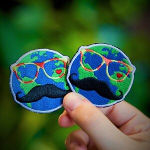 May include: Two embroidered patches shaped like the Earth, each with a black mustache, red heart-shaped eyes, and blue-rimmed glasses. The patches are held in a hand against a green background.
