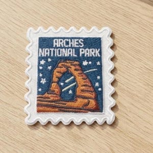 May include: A rectangular embroidered patch shaped like a postage stamp with a white scalloped border. The design features a dark blue night sky with white stars and a brown rock arch formation with the text Arches National Park in white.