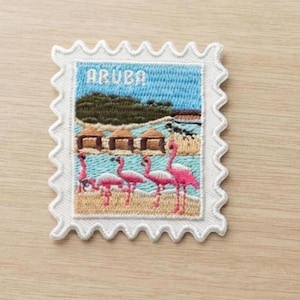 May include: A rectangular embroidered patch shaped like a postage stamp with a white scalloped border. The design features a beach scene with pink flamingos, tan huts, a green hill, and a blue sky, with the word ARUBA stitched in white letters.