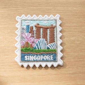 Op de afbeelding: Een rechthoekige geborduurde patch in de vorm van een postzegel met een geschulpte witte rand. Het ontwerp toont een kleurrijke afbeelding van de skyline van Singapore, inclusief het Marina Bay Sands, met het woord SINGAPORE in wit geborduurd.
