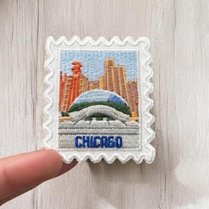 Könnte beinhalten: Ein rechteckiger, bestickter Aufnäher in Form einer Briefmarke mit gewelltem weißem Rand. Das Design zeigt die Skyline von Chicago mit hohen Gebäuden und einer markanten Kuppelstruktur sowie dem Wort CHICAGO in blauer Schrift darunter.