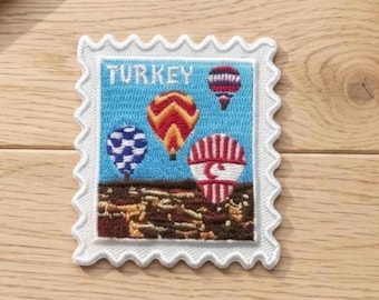 May include: A rectangular embroidered patch shaped like a postage stamp with a scalloped white border. The design features four colorful hot air balloons floating over a brown landscape against a blue sky, with the word TURKEY printed in white.