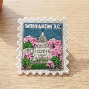 May include: A rectangular embroidered patch shaped like a postage stamp featuring the United States Capitol building in Washington D.C. The design includes pink cherry blossoms, green trees, and a blue sky, with the text WASHINGTON D.C. at the top.