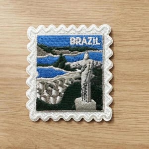 May include: A square embroidered patch with a scalloped white border featuring a landscape of Brazil. The design depicts the Christ the Redeemer statue in white against a dark green mountain, blue water, and a light blue sky with the word BRAZIL.