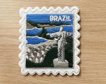 May include: A square embroidered patch with a scalloped white border featuring a landscape of Brazil. The design depicts the Christ the Redeemer statue in white against a dark green mountain, blue water, and a light blue sky with the word BRAZIL.