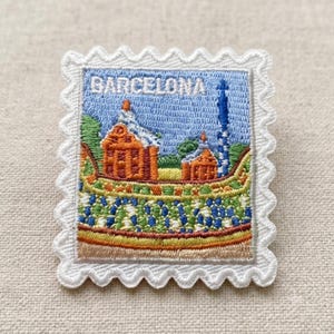 Peut inclure: Un écusson brodé rectangulaire en forme de timbre-poste avec une bordure blanche festonnée. Le motif présente le texte BARCELONA en blanc, avec des bâtiments orange, une tour bleue et un motif en mosaïque coloré sur un ciel bleu clair.