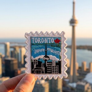 May include: A rectangular embroidered patch shaped like a postage stamp with a white scalloped border. The design features the word TORONTO in blue letters, a red maple leaf, and a black silhouette of the city skyline including the CN Tower.