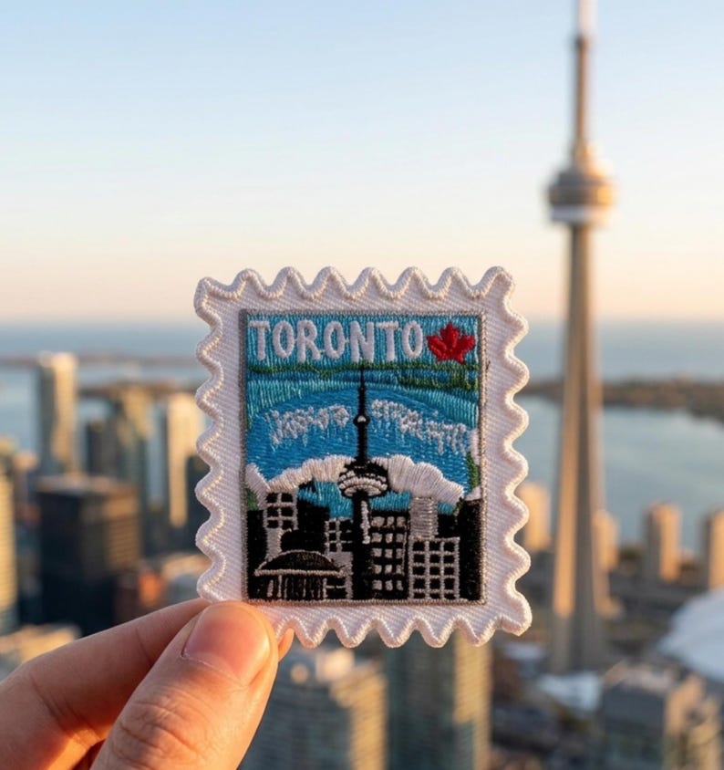 May include: A rectangular embroidered patch shaped like a postage stamp with a white scalloped border. The design features the word TORONTO in blue letters, a red maple leaf, and a black silhouette of the city skyline including the CN Tower.