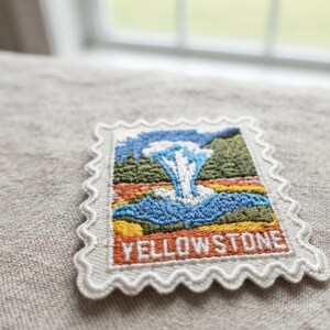 May include: A rectangular embroidered patch shaped like a postage stamp with a scalloped border. The design features a blue geyser erupting against a green forest and orange landscape, with the word YELLOWSTONE stitched in white at the bottom.