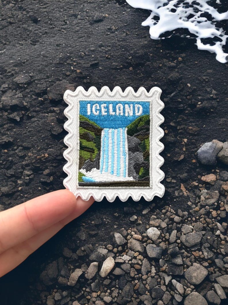 May include: A rectangular embroidered patch with a scalloped white border, featuring a landscape illustration of a waterfall between green hills and rocky terrain, with the word ICELAND in white capital letters against a blue sky background.