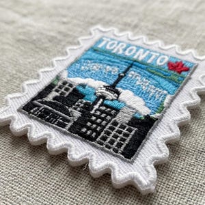 May include: A rectangular embroidered patch shaped like a postage stamp with a white scalloped border. The design features the Toronto skyline in black thread against a light blue sky, with a red maple leaf and the word TORONTO in white.