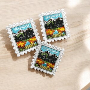 Puede incluir: Tres parches bordados rectangulares con forma de sello postal y bordes blancos festoneados. Cada parche muestra un paisaje de Vancouver con monta&ntilde;as nevadas, cielo azul, una masa de agua y &aacute;rboles de oto&ntilde;o en tonos naranja y amarillo.