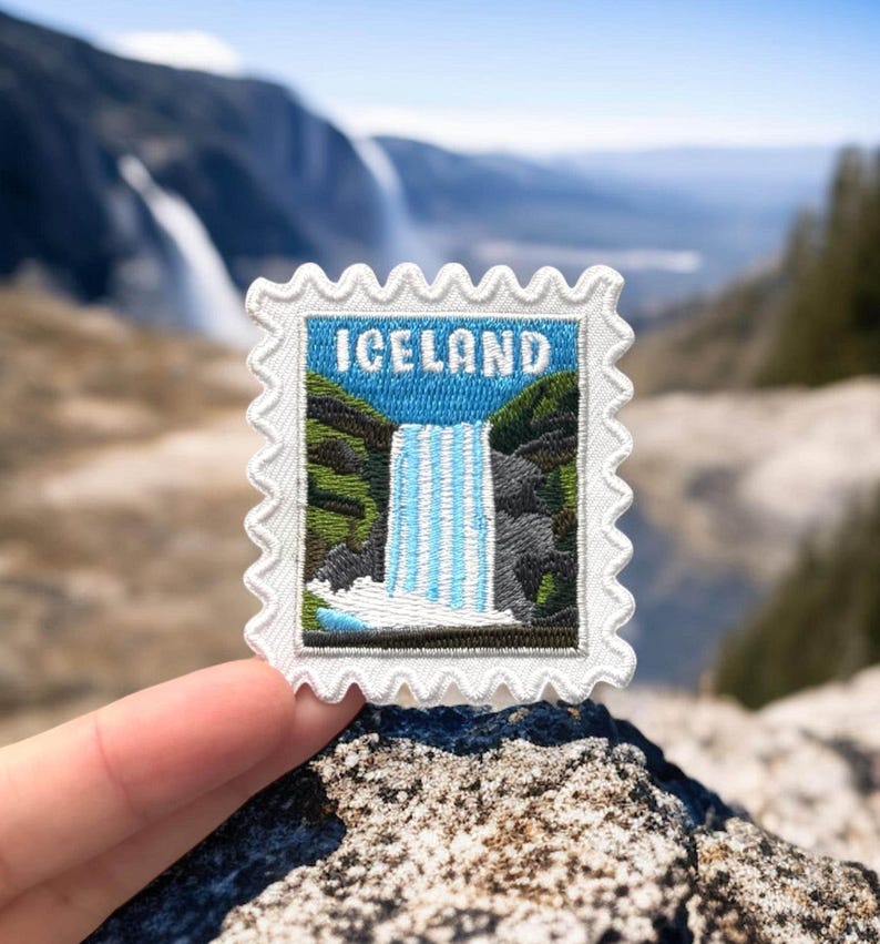 May include: A white embroidered patch in the shape of a postage stamp, featuring a waterfall scene and the word "ICELAND" in blue. The patch is held against a blurred natural background of mountains and sky.