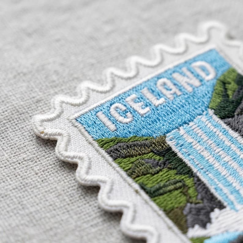 May include: A rectangular embroidered patch shaped like a postage stamp with a scalloped white border. The design features the word ICELAND in white letters against a light blue sky, with green hills and a cascading white waterfall below.