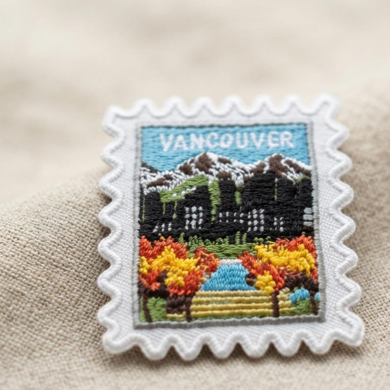 Puede incluir: Un parche bordado rectangular con forma de sello postal que incluye la palabra VANCOUVER en letras blancas. El dise&ntilde;o muestra un horizonte urbano con edificios negros, monta&ntilde;as nevadas, un cielo azul y &aacute;rboles oto&ntilde;ales coloridos.
