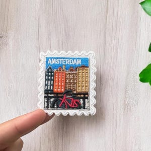 May include: A square embroidered patch with a scalloped white border featuring a blue sky, a row of colorful buildings in black, orange, brown, and tan, and a red bicycle in the foreground. The text AMSTERDAM is stitched in white at the top.